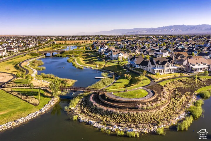 Aerial perspective of a community featuring a large water element, multi-tiered landscaped embankments with stone retaining walls, paved pedestrian pathways, and steel footbridges, set against a backdrop of mountain ranges
