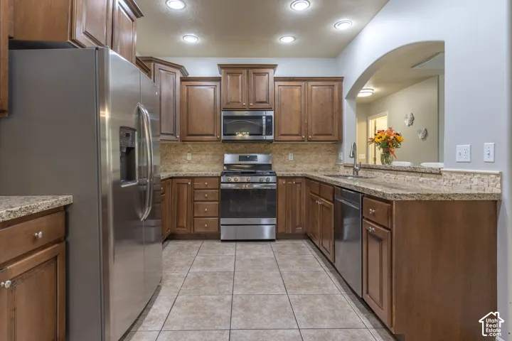 Kitchen featuring wood cabinetry, stone-finish countertops, stainless steel appliances, tiled flooring, and an arched wall opening