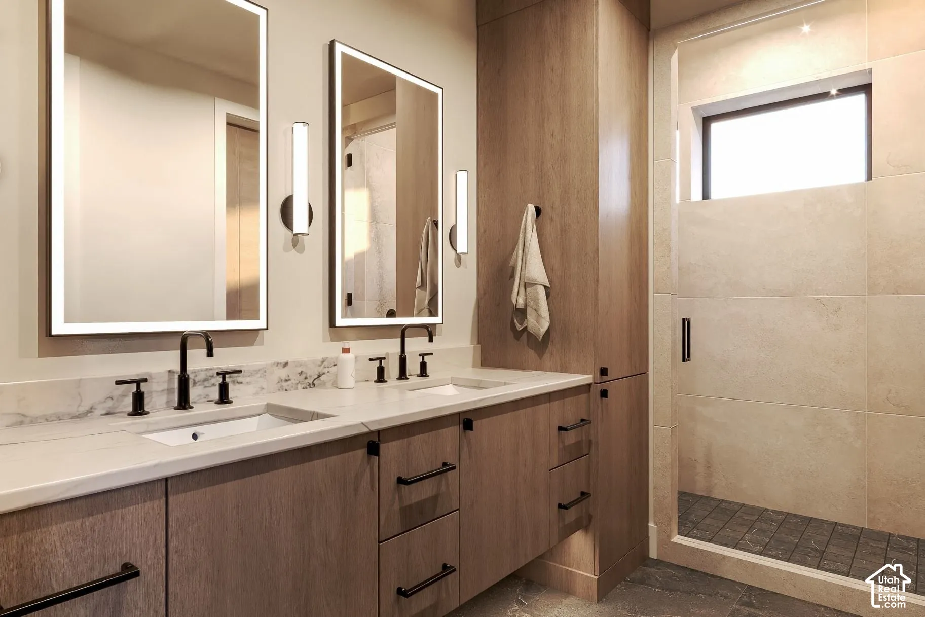 Dual-sink vanity with stone countertop, wood-finish cabinetry, backlit vanity mirrors, matte black fixtures, and a walk-in shower with stone tile surround