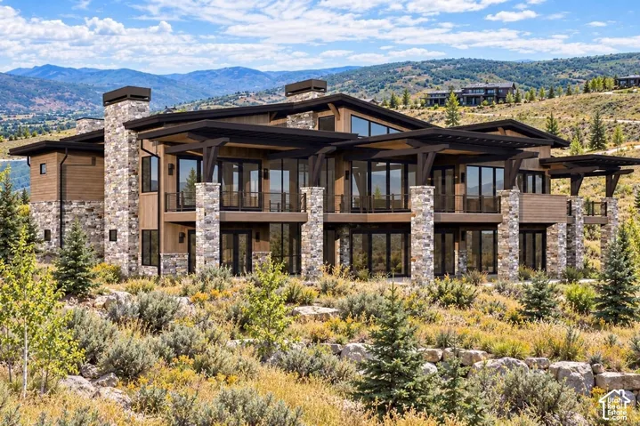 Modern multi-level exterior featuring stacked stone masonry, horizontal wood siding, floor-to-ceiling windows, deep covered balconies, and expansive mountain terrain