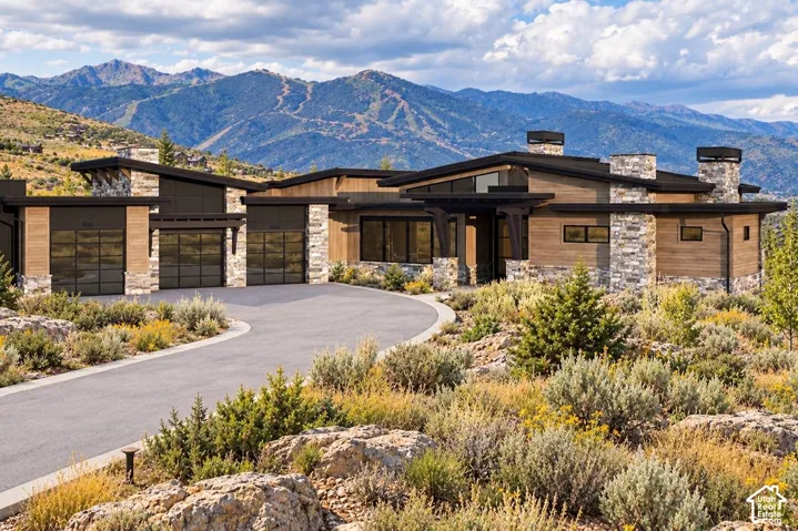 Modern mountain-style residence featuring horizontal wood siding, stone masonry chimneys, shed rooflines, oversized multi-bay garage doors, and mountain range backdrop