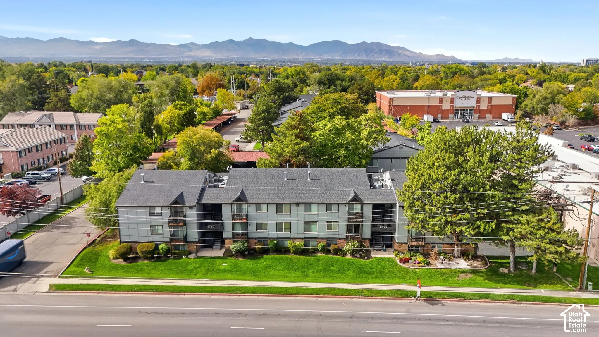 Multi-story residential building featuring shingled roof lines, exterior balconies, landscaped lawns, mature trees, and mountain backdrop