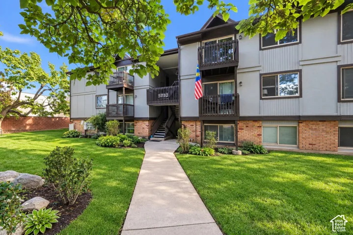 Multi-level exterior featuring a brick and siding facade, private balconies, and concrete walkway leading to the building entrance, surrounded by landscaped grounds