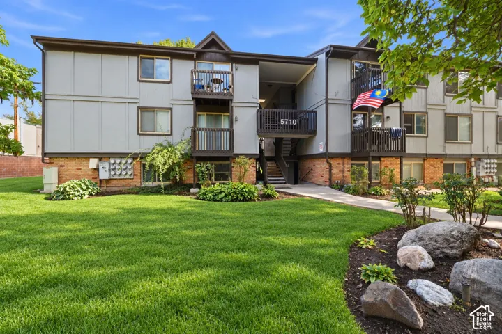 Multi-level exterior featuring light-toned paneling, brick masonry, private balconies, and concrete walkways surrounded by landscaped grounds