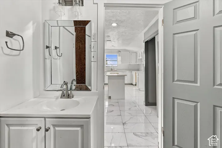 Bathroom featuring a white vanity with an integrated basin, a rectangular framed mirror, and dual-handle metal faucet, with light-toned stone-finish tile flooring extending into the kitchen
