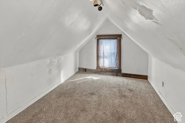 Vaulted ceiling featuring a central window with rustic wood trim, neutral wall-to-wall carpeting, and surface-mounted ceiling track lighting