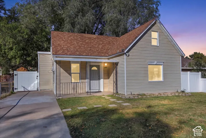 Exterior featuring light-toned horizontal siding, a covered front porch with iron railing, a concrete driveway, and white vinyl perimeter fencing