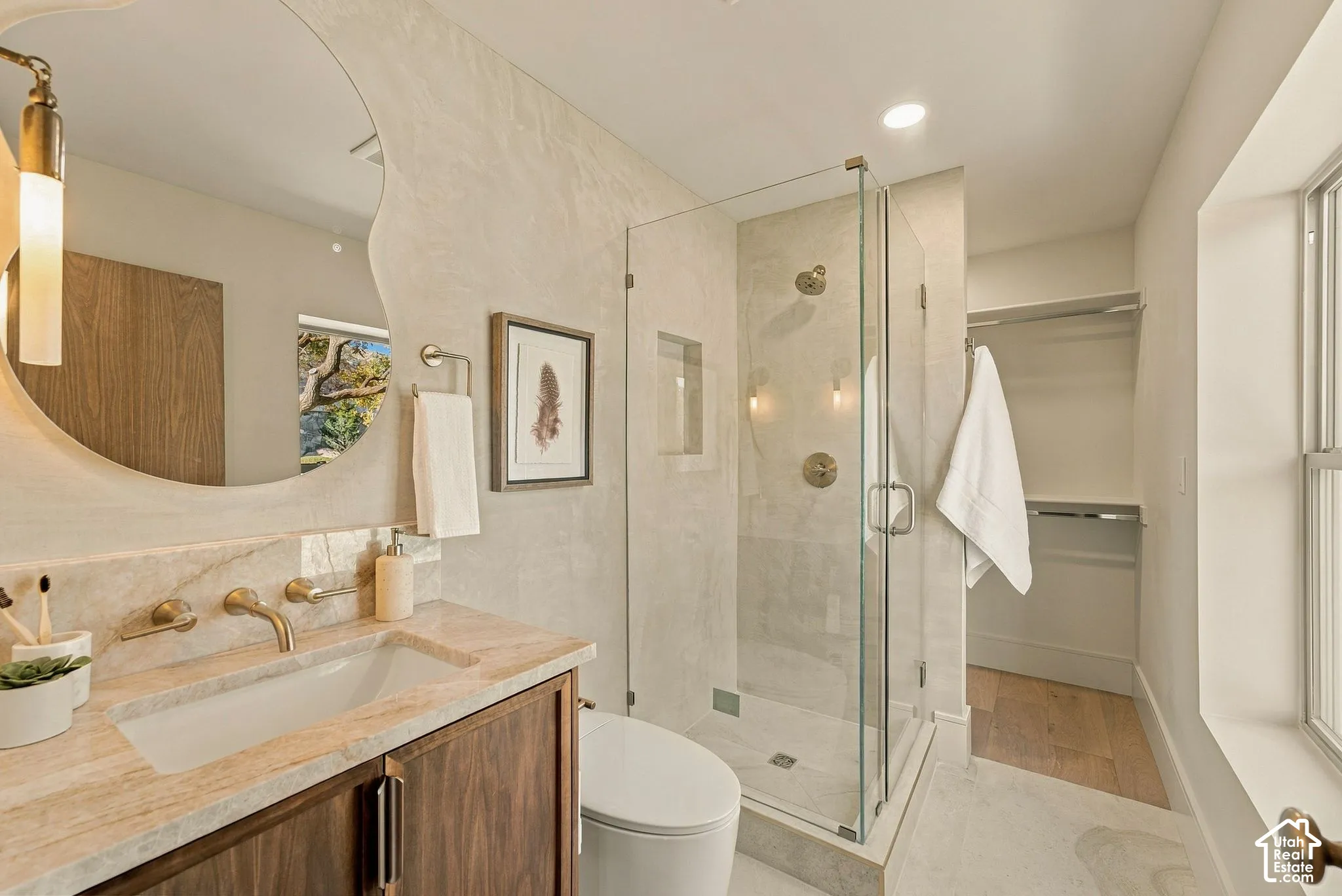 Bathroom featuring a wood-finish vanity with a stone countertop, an irregular-shaped wall mirror, a glass-enclosed shower with dual showerheads, and recessed wall shelving