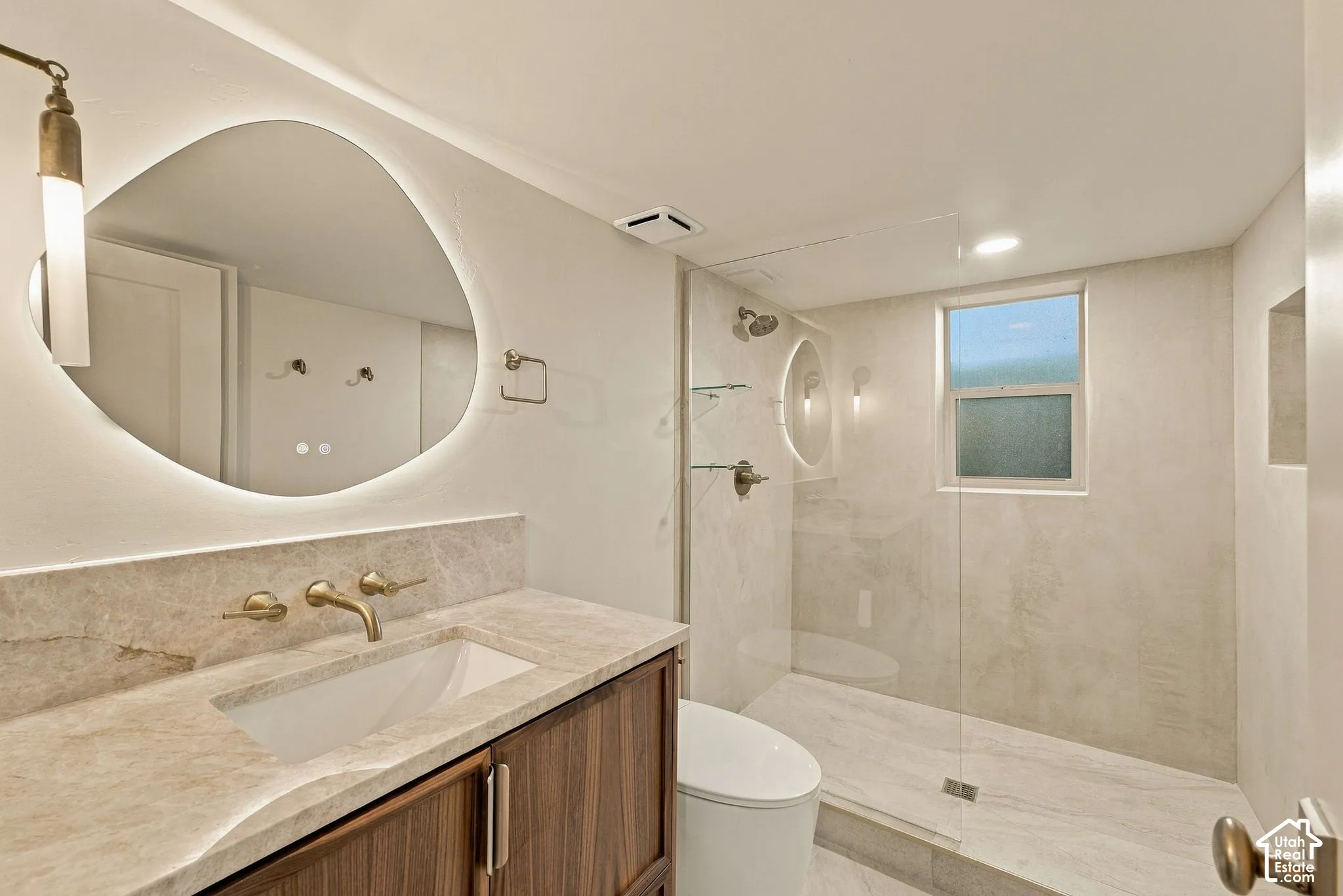 Bathroom featuring a custom wood-finish vanity with a stone countertop, an illuminated organic-shaped mirror, and a walk-in shower with a glass enclosure and wall-mounted hardware