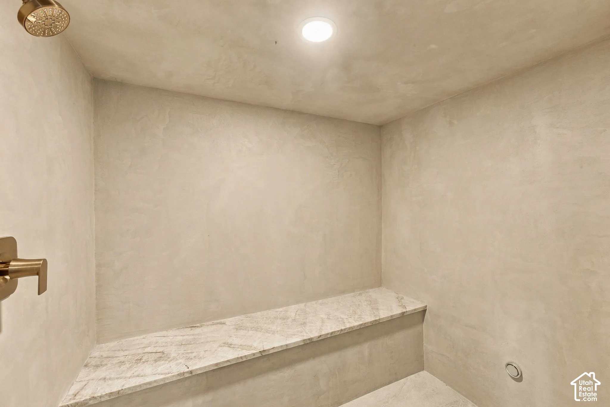 Walk-in shower enclosure with a stone-top built-in bench, seamless plaster walls, gold-finish faucet hardware, and recessed ceiling lighting