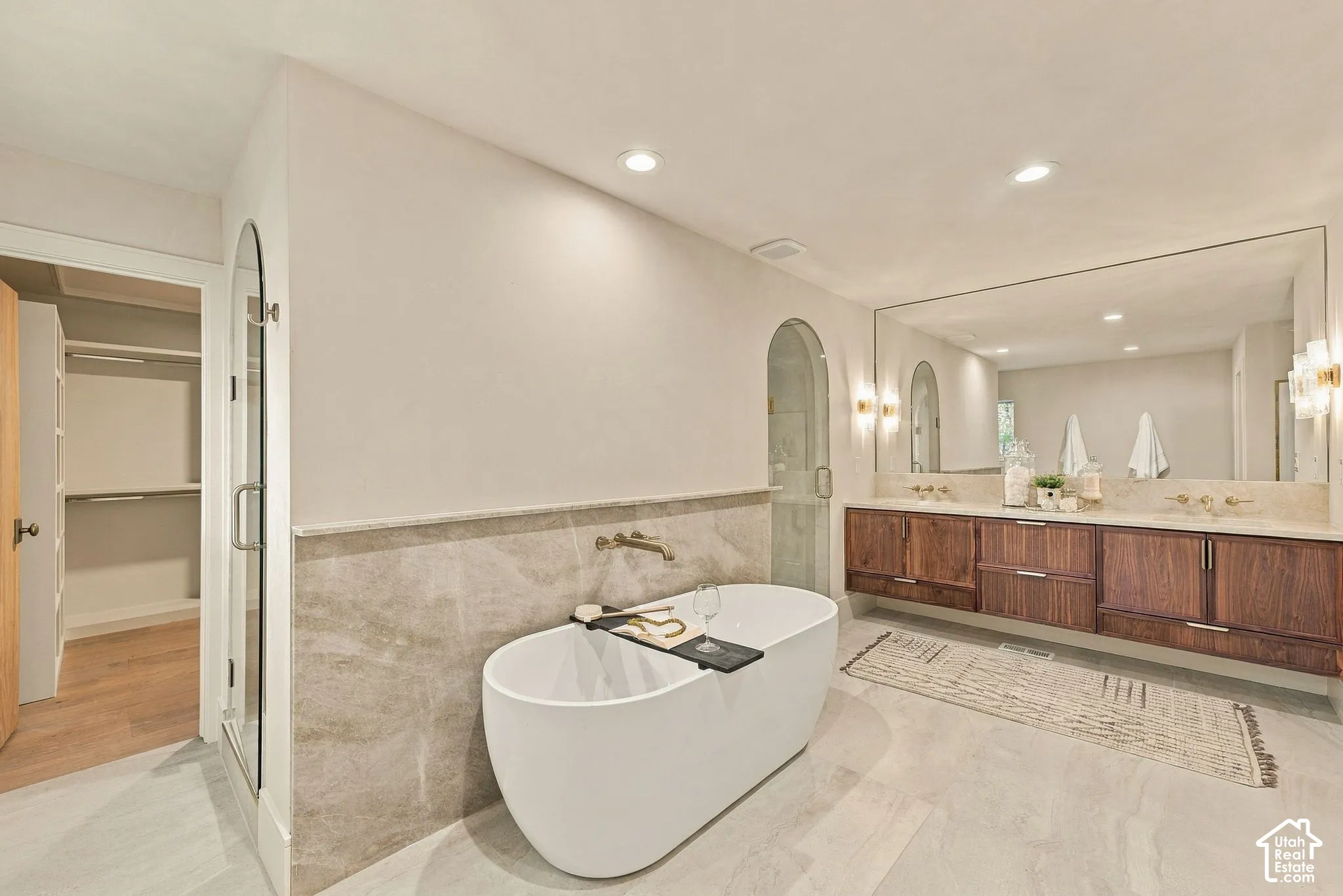Freestanding soaking tub with wall-mounted faucet, stone tile wainscoting, floating wood-finish vanity with dual sinks, recessed lighting, and expansive wall mirror