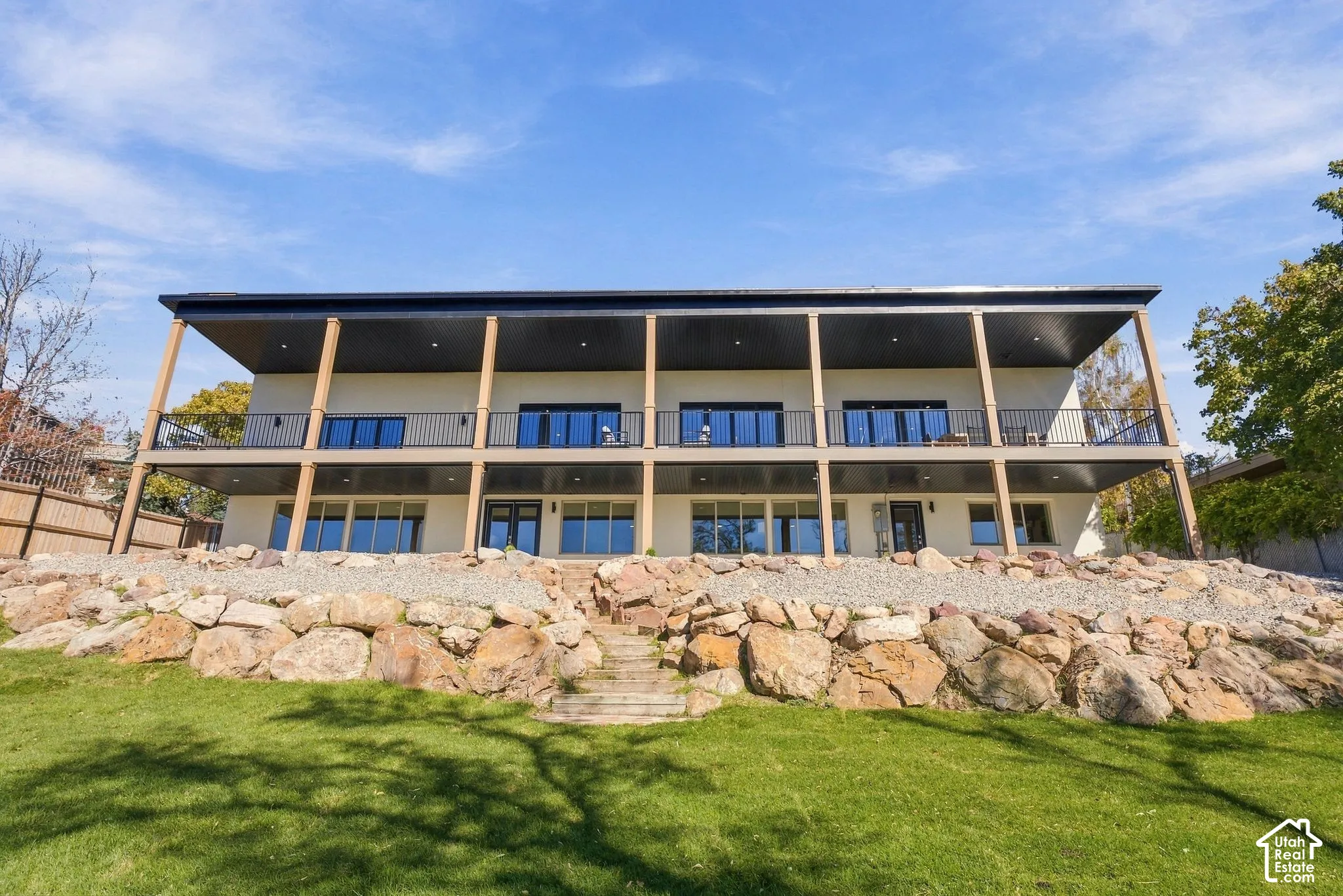 Two-story exterior with dual-level covered balconies, metal railing, recessed lighting, and expansive stone retaining wall