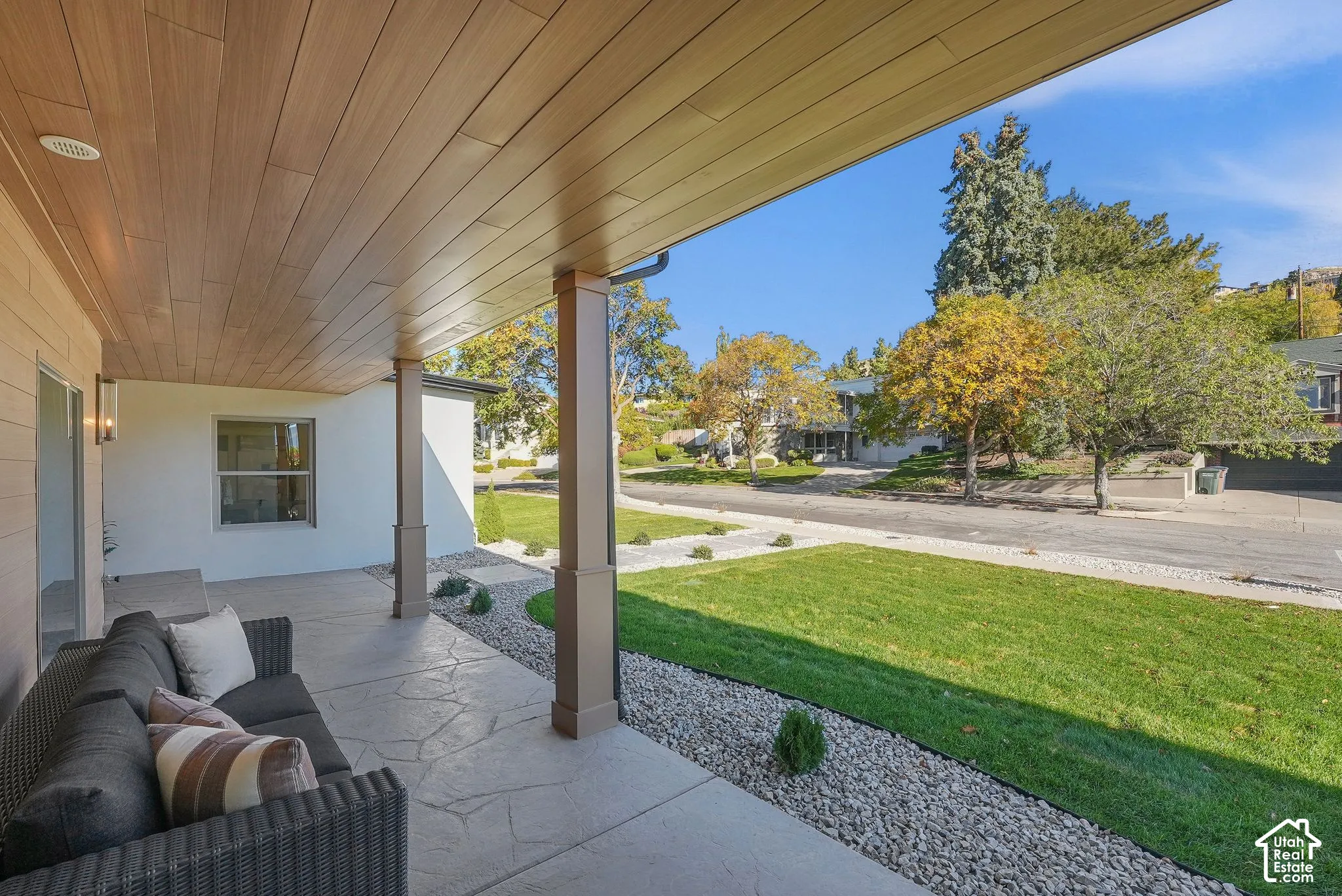 Covered patio featuring a wood-finished tongue-and-groove ceiling, stone-patterned concrete flooring, and square support columns, overlooking a manicured lawn with gravel landscaping borders