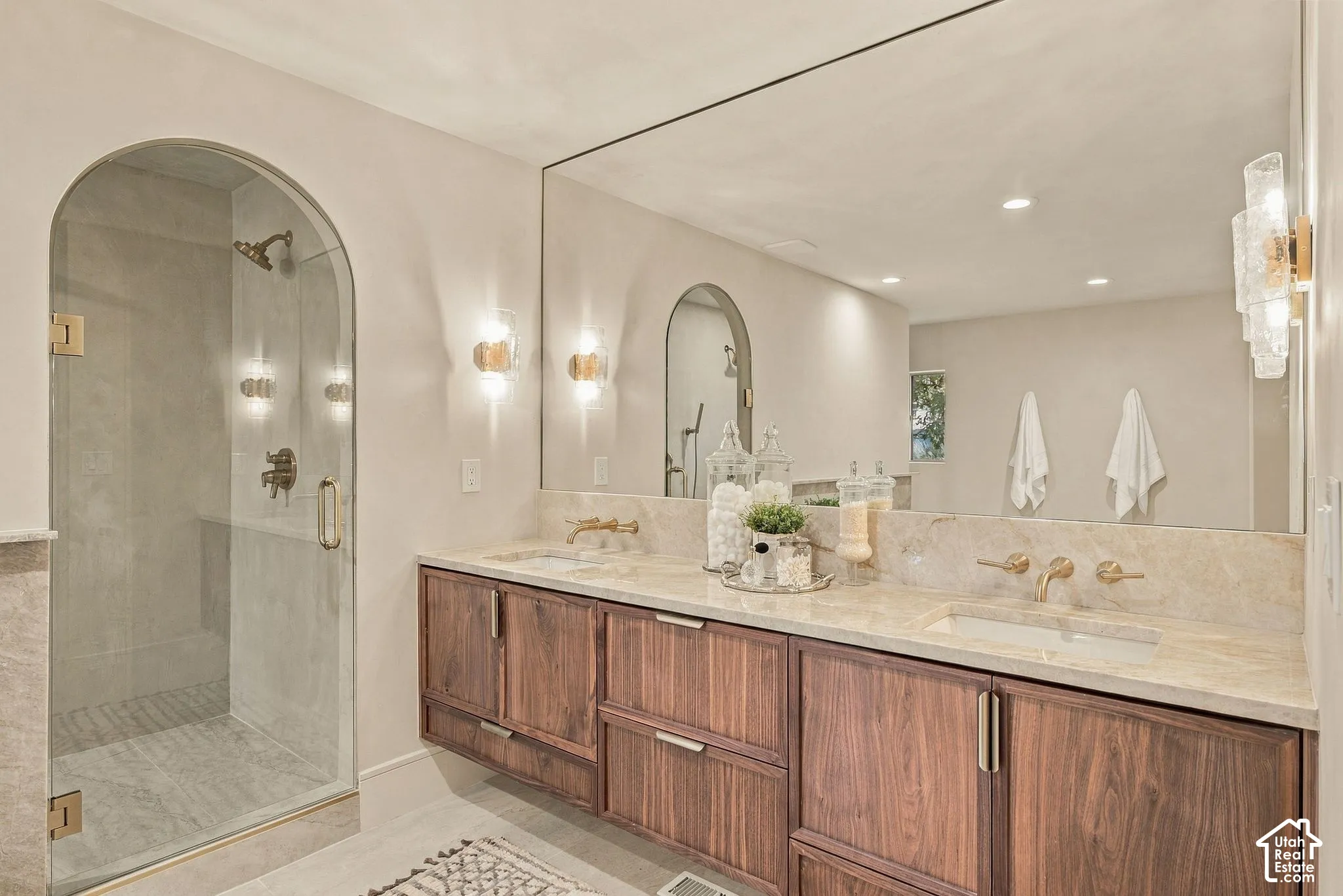 Floating wood-finish vanity with dual integrated sinks, stone countertop, arched glass-enclosed shower with gold-tone hardware, and expansive wall-mounted mirror