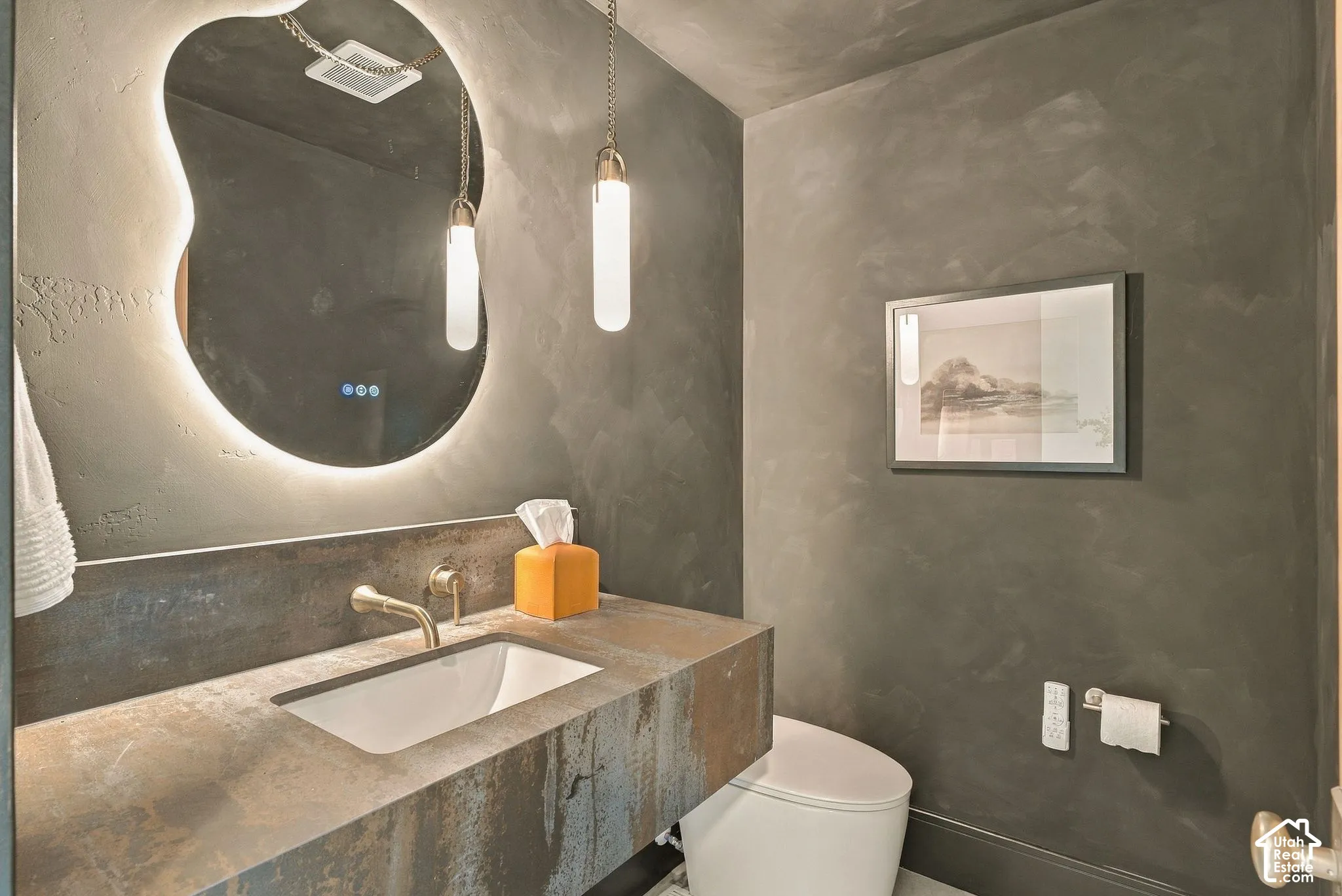Custom vanity featuring a textured finish and integrated sink, illuminated wall-mounted mirror, brushed metal wall-mounted faucet, and pendant lighting