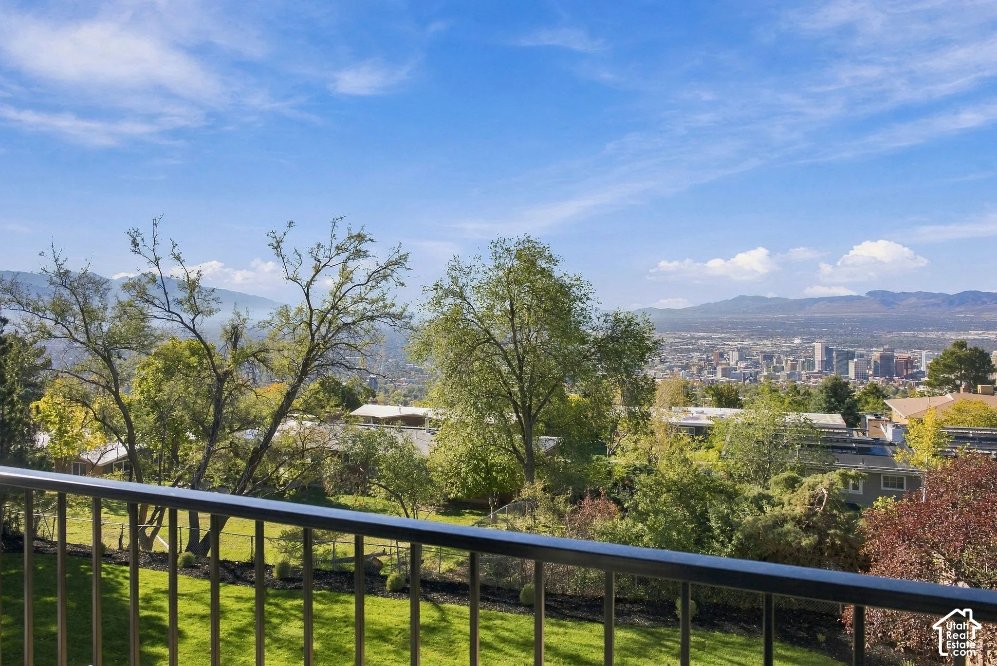 Elevated balcony featuring a matte-finish metal railing, overlooking an expansive landscape of mature trees, manicured lawn, and a mountain and city skyline backdrop