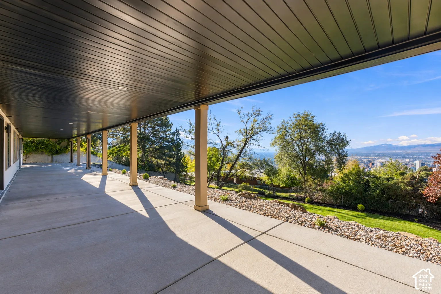 Covered concrete patio featuring a dark-toned tongue-and-groove ceiling, recessed lighting, and structural support pillars overlooking the natural landscape