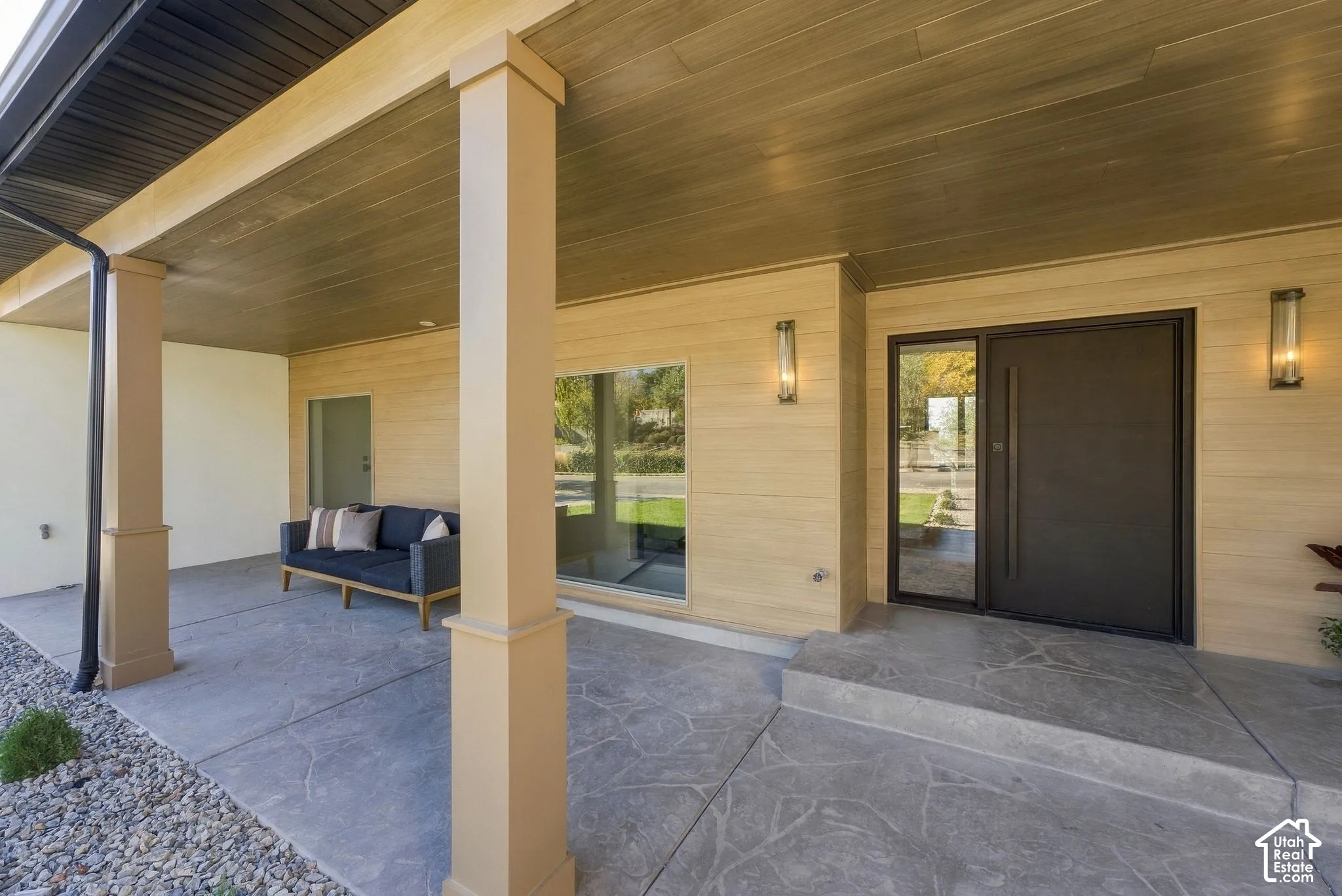 Covered entryway featuring horizontal wood-tone siding, decorative stone-patterned concrete flooring, an oversized solid entry door with a vertical pull handle, and structured square support columns