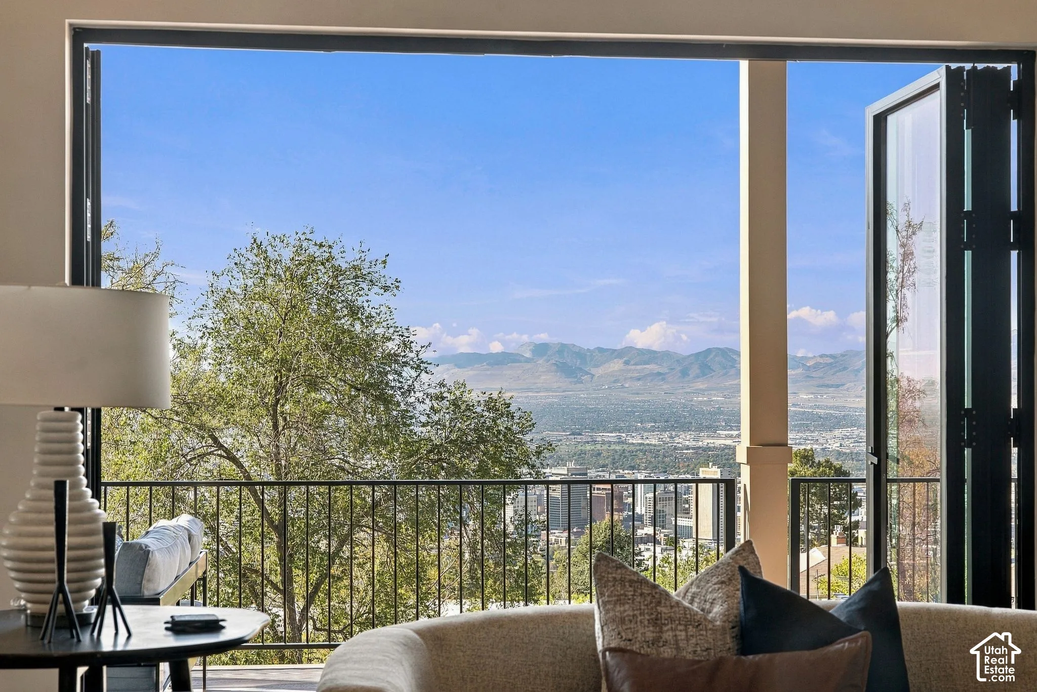 Floor-to-ceiling bi-fold glass doors opening to a balcony with metal railing, featuring a panoramic mountain and city backdrop
