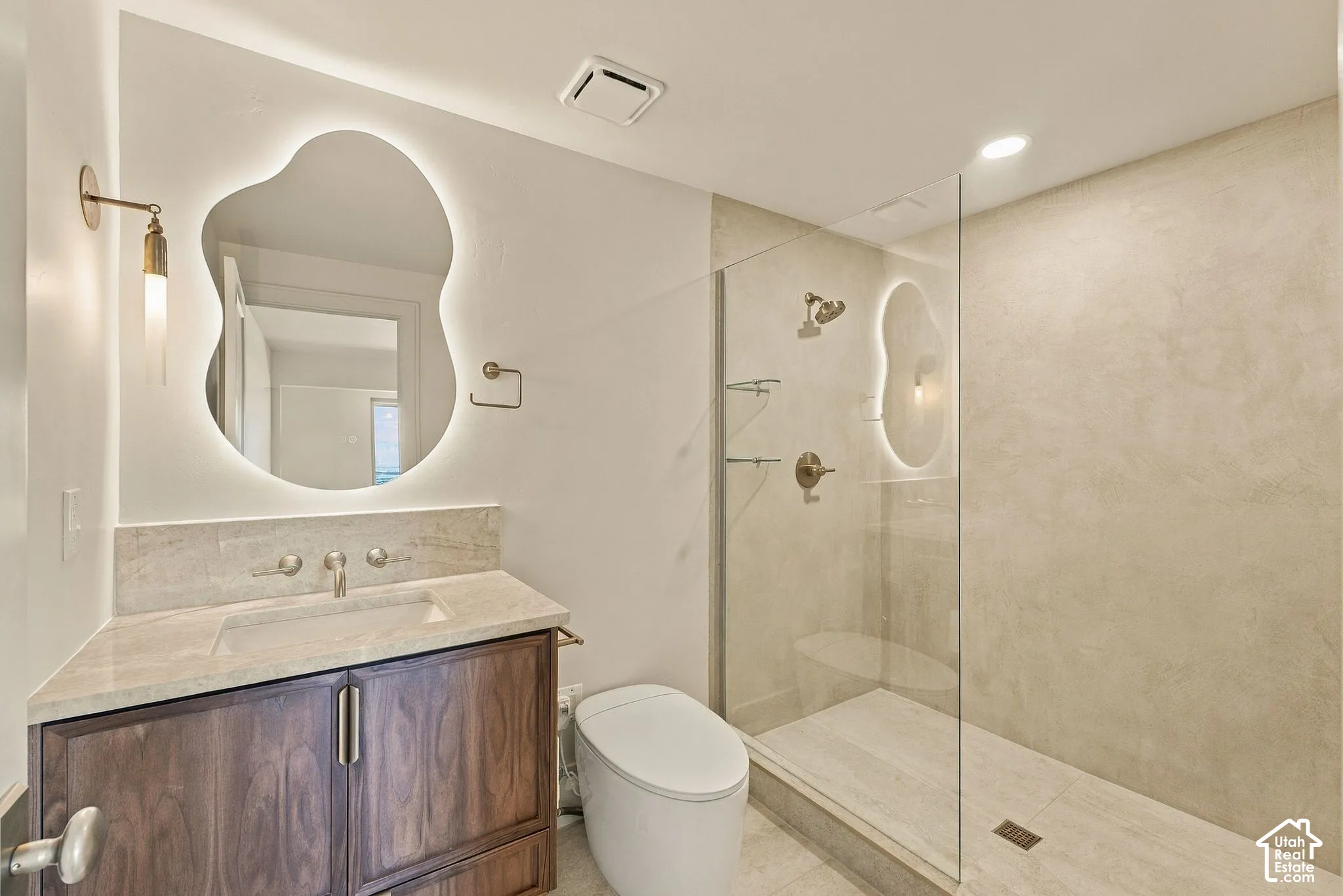 Bathroom featuring wood-finish cabinetry, stone countertop, wall-mounted faucet, asymmetrical backlit mirror, and a walk-in shower with a glass panel and textured tile surround