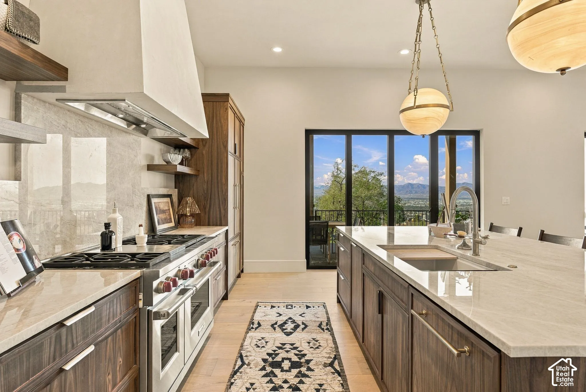 Gourmet kitchen featuring a large central island with light-toned stone countertops, custom wood-finish cabinetry, a professional-grade gas range with a sleek vent hood, and multi-light globe pendant fixtures