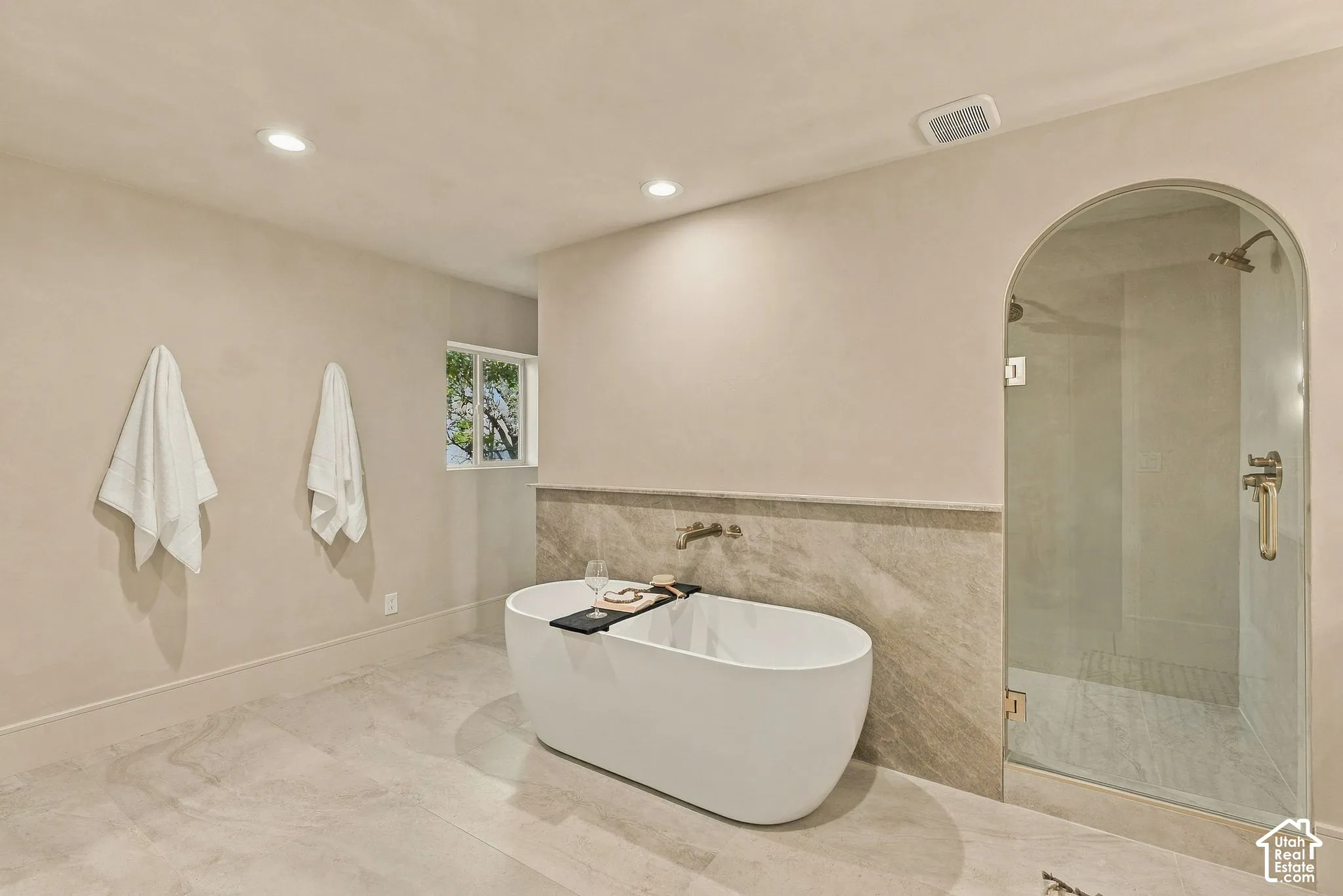 Freestanding oval soaking tub with wall-mounted faucet, arched glass-enclosed shower, stone-finish tile flooring, and stone-tiled wainscoting