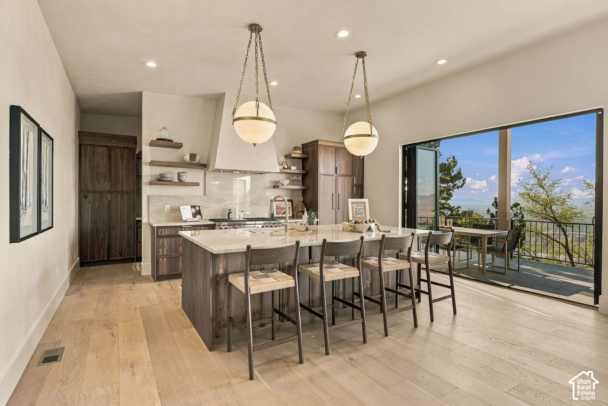 Kitchen featuring a large central island with quartz countertops, custom dark wood cabinetry, floating wall shelves, a professional-style range hood, and floor-to-ceiling bifold doors opening to an outdoor patio