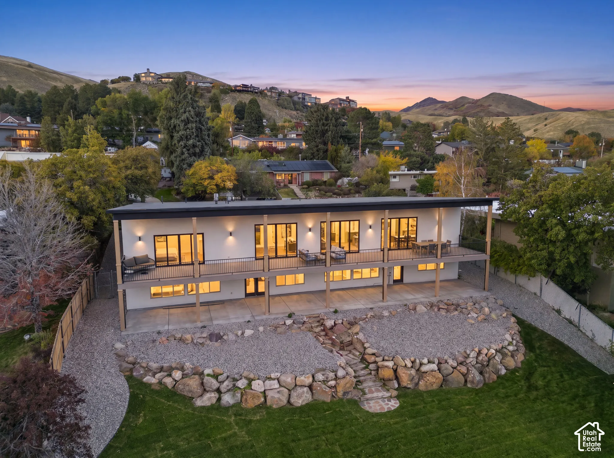 Contemporary two-story exterior featuring multi-panel sliding glass doors, a full-length elevated balcony with metal railings, a covered lower patio, and a tiered rock garden with natural stone masonry