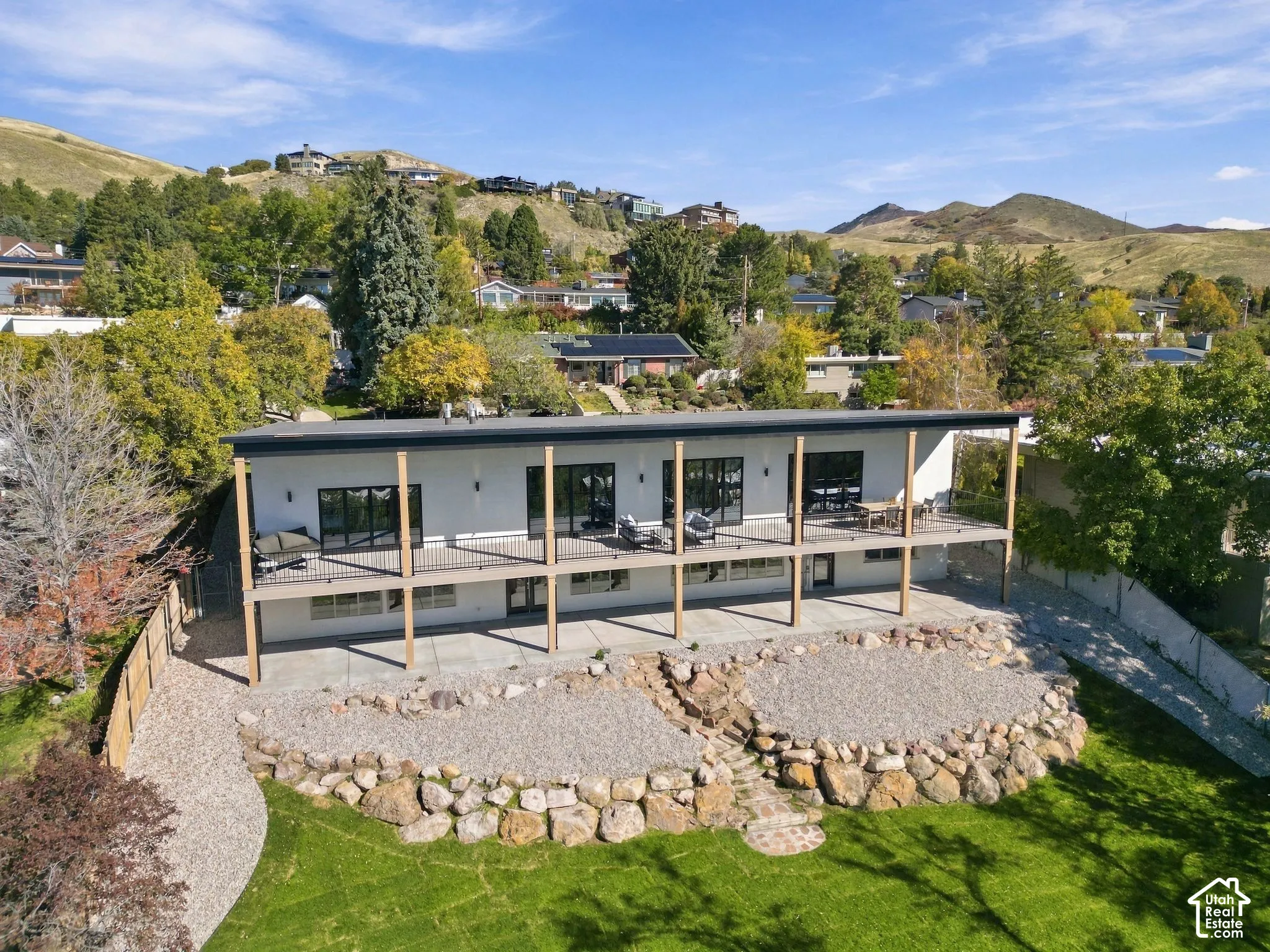 Contemporary multi-level exterior featuring an expansive wrap-around deck, integrated structural support columns, floor-to-ceiling glass sliding doors, stone hardscaping, and a terraced landscape design