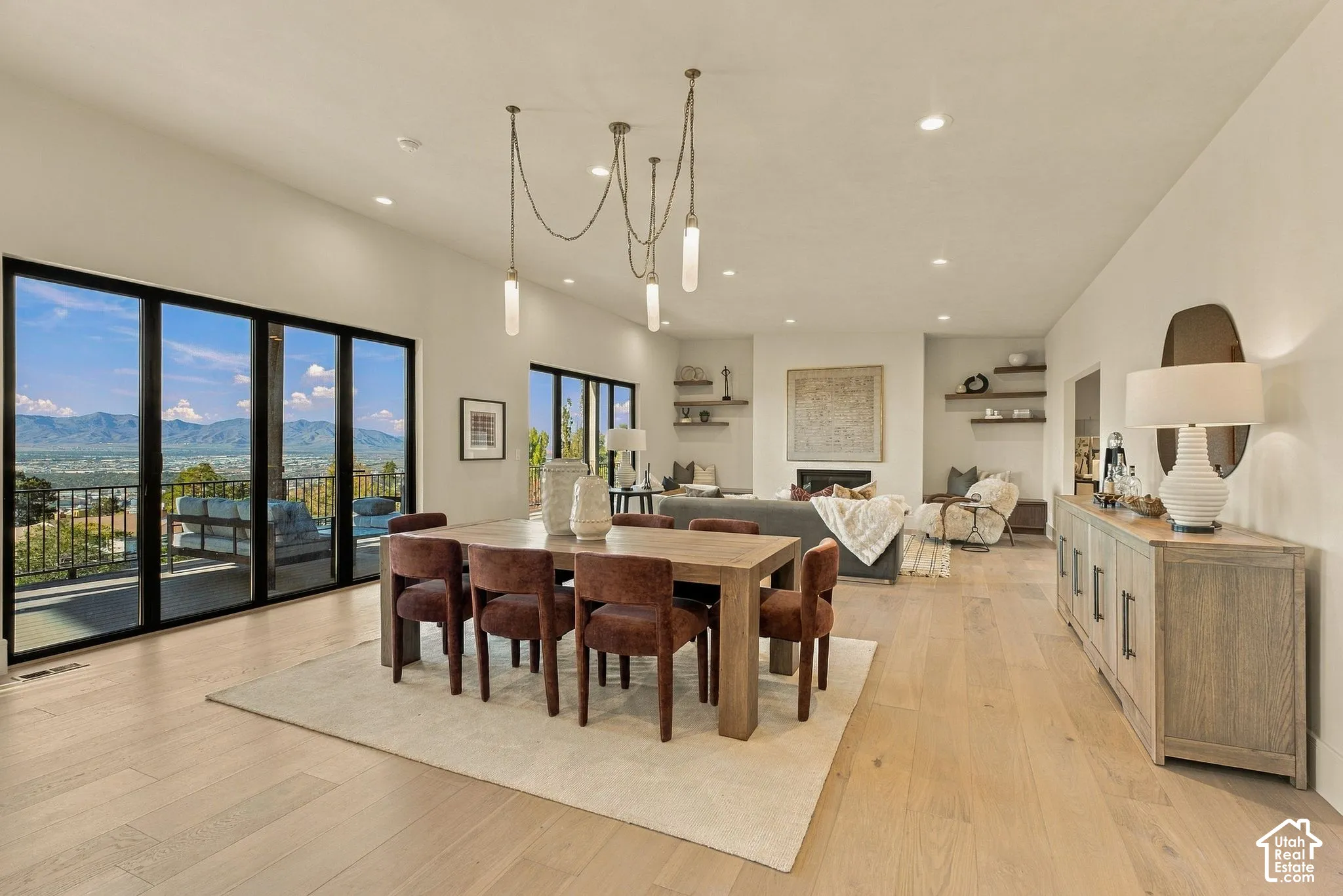 Open-concept room featuring wood-finish flooring, recessed lighting, multi-panel sliding glass doors with mountain scenery, built-in floating shelves, and a multi-pendant light fixture