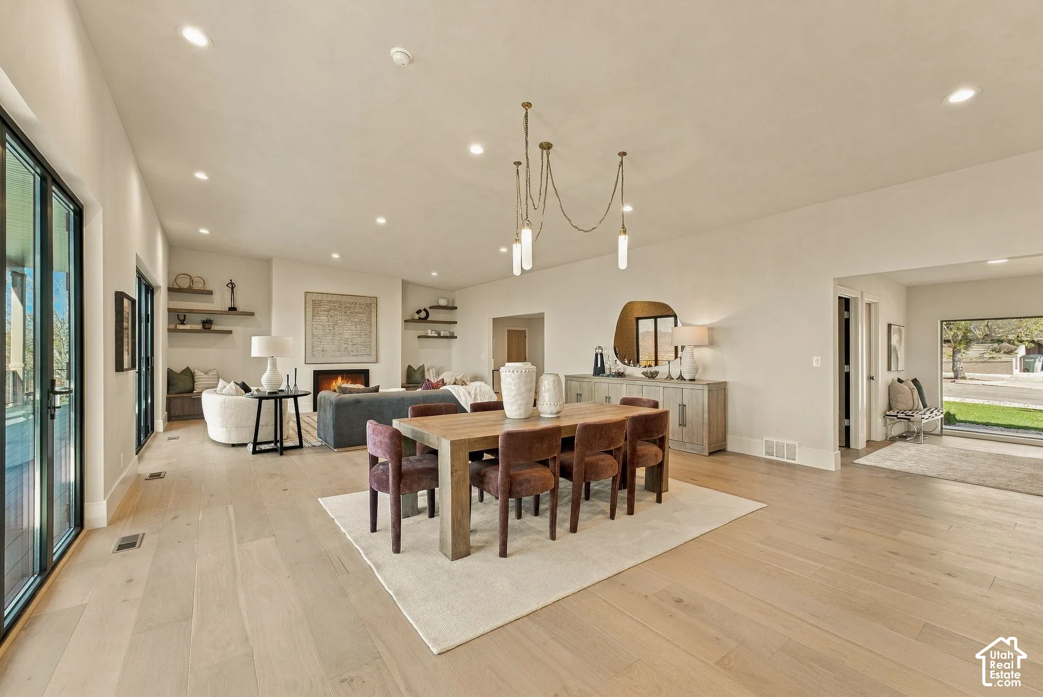 Open-concept interior featuring wide-plank wood-finish flooring, a contemporary multi-arm chandelier, built-in wall shelving, a fireplace with a minimalist surround, and expansive floor-to-ceiling multi-panel glass doors