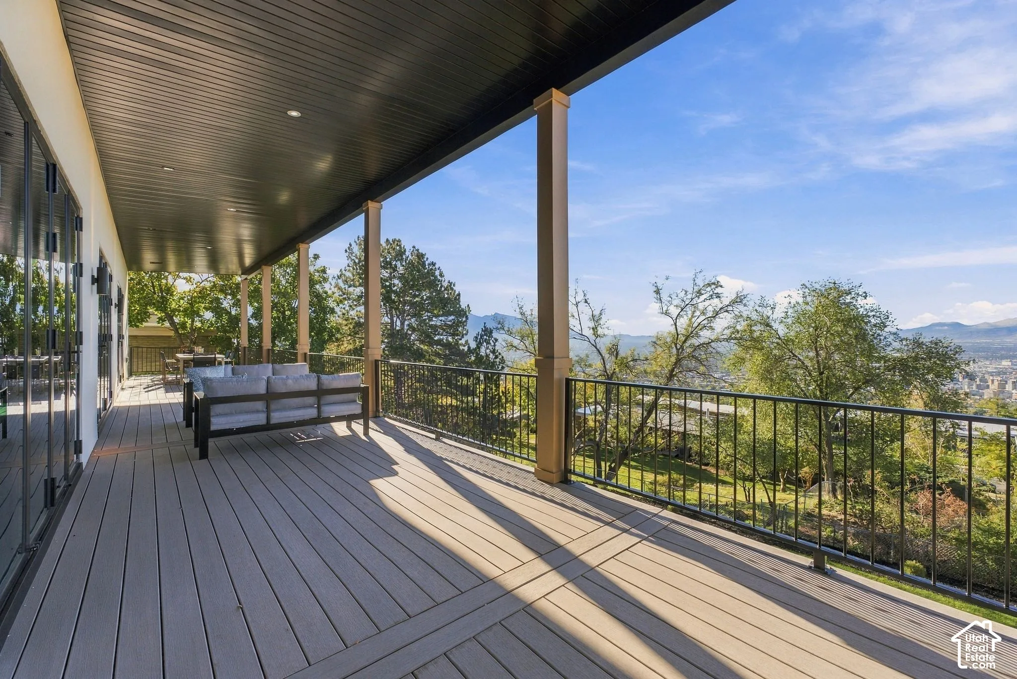 Covered balcony featuring composite decking, recessed lighting, square columns, and a black metal railing overlooking the surrounding landscape