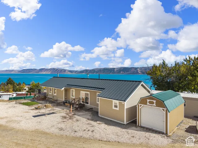 Exterior featuring tan horizontal siding, a dark metal roof, a detached storage building with a roll-up door, and mountain and water scenery