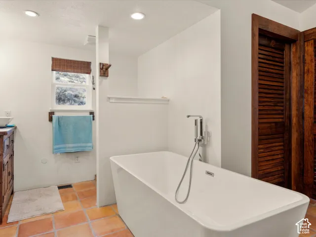 Freestanding soaking tub with a chrome handheld faucet, natural clay tile flooring, built-in wooden louvered closet doors, and recessed ceiling lighting