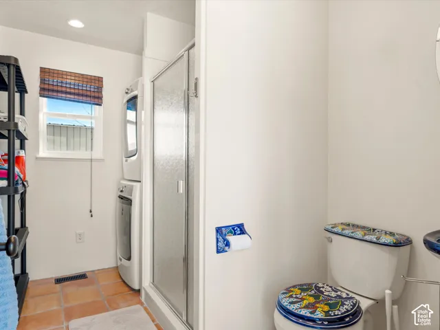 Stacked laundry unit, shower with glass enclosure, terra-cotta tile flooring, and single-hung window with a woven shade