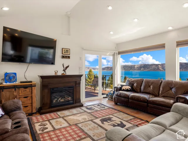 Vaulted ceilings with recessed lighting, dark-wood fireplace surround, large picture windows, and sliding glass door with access to an exterior deck featuring a mountainous lake backdrop