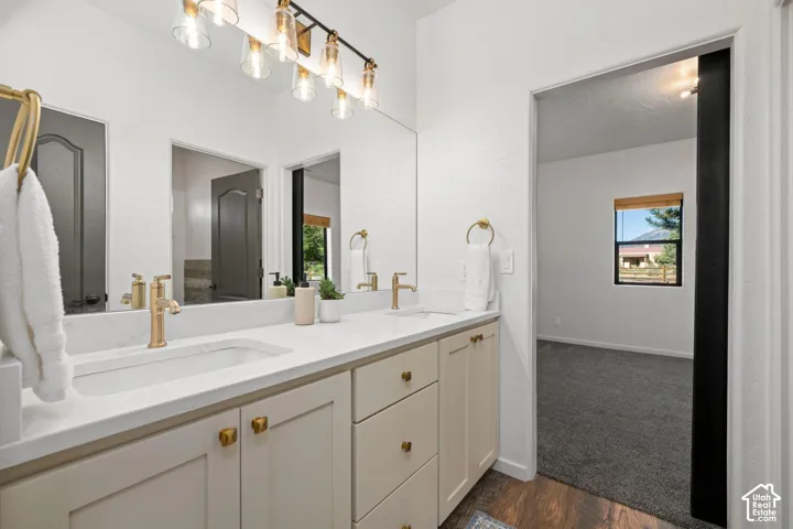Bathroom featuring a white countertop with integrated sinks, brass-toned fixtures, light-toned cabinetry with brass-toned hardware, and an expansive wall-to-wall mirror with multi-bulb light bar