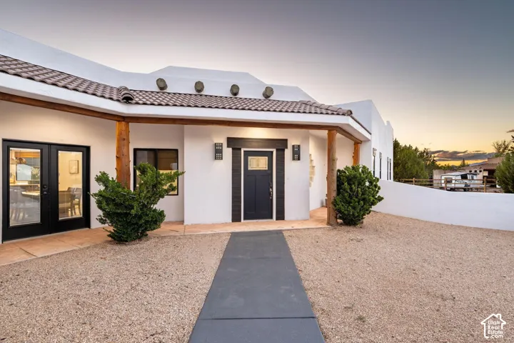 Pueblo-style exterior featuring a covered porch with exposed log columns, terracotta tile roof detailing, smooth stucco finish, and an inset entry door with modern geometric wall sconces
