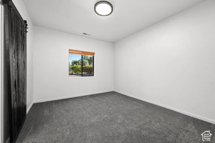 Interior room featuring charcoal-toned carpeting, white walls, a dark-finish sliding barn door, and a single-pane window with wood-finish blinds