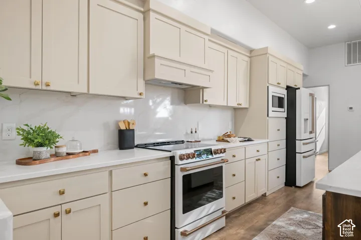 Kitchen featuring custom shaker-style cabinetry, solid-surface countertops, full-height stone-look backsplash, wood-finish flooring, and integrated white appliances with metallic hardware