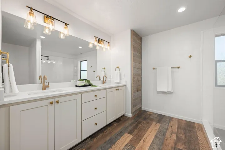 Double vanity with white cabinetry, light-toned countertop, integrated sinks, brass-finish fixtures, and full-width vanity mirror with integrated multi-light sconces, accented by wood-finish flooring and a vertical wood-plank wall feature