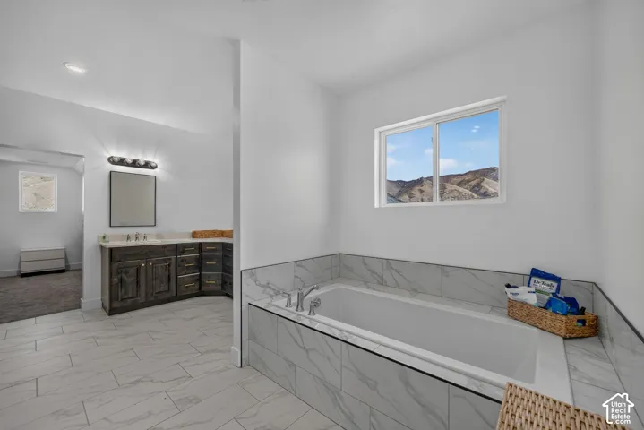 Soaking tub featuring large-format tile surround, dual-sink vanity with stone countertop, tile flooring, and mountain backdrop
