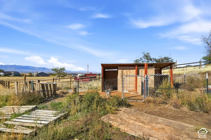 Outbuilding with an open-sided shelter and wire-mesh fencing, set within a rural landscape featuring mountain backdrops and post-and-rail perimeter fencing