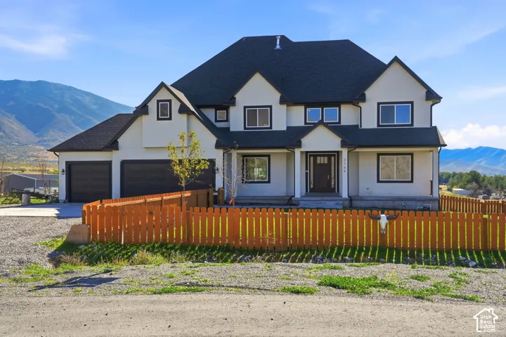 Two-story residence featuring a stucco exterior, multi-gabled asphalt shingle roof, attached three-car garage, and a wood picket fence, set against a mountain backdrop