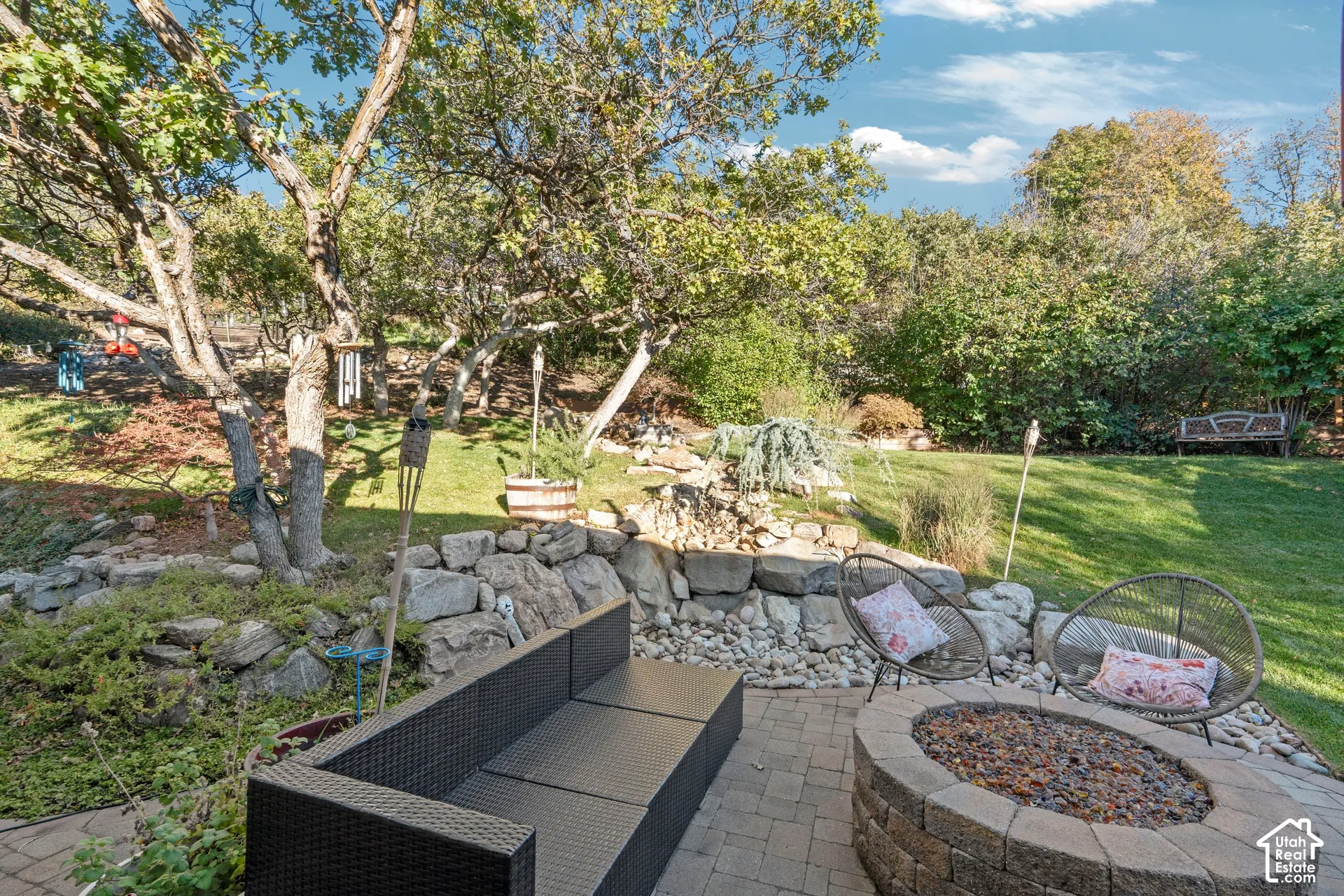 Backyard featuring a paved stone patio, built-in circular fire pit, natural stone retaining wall, and mature landscaping