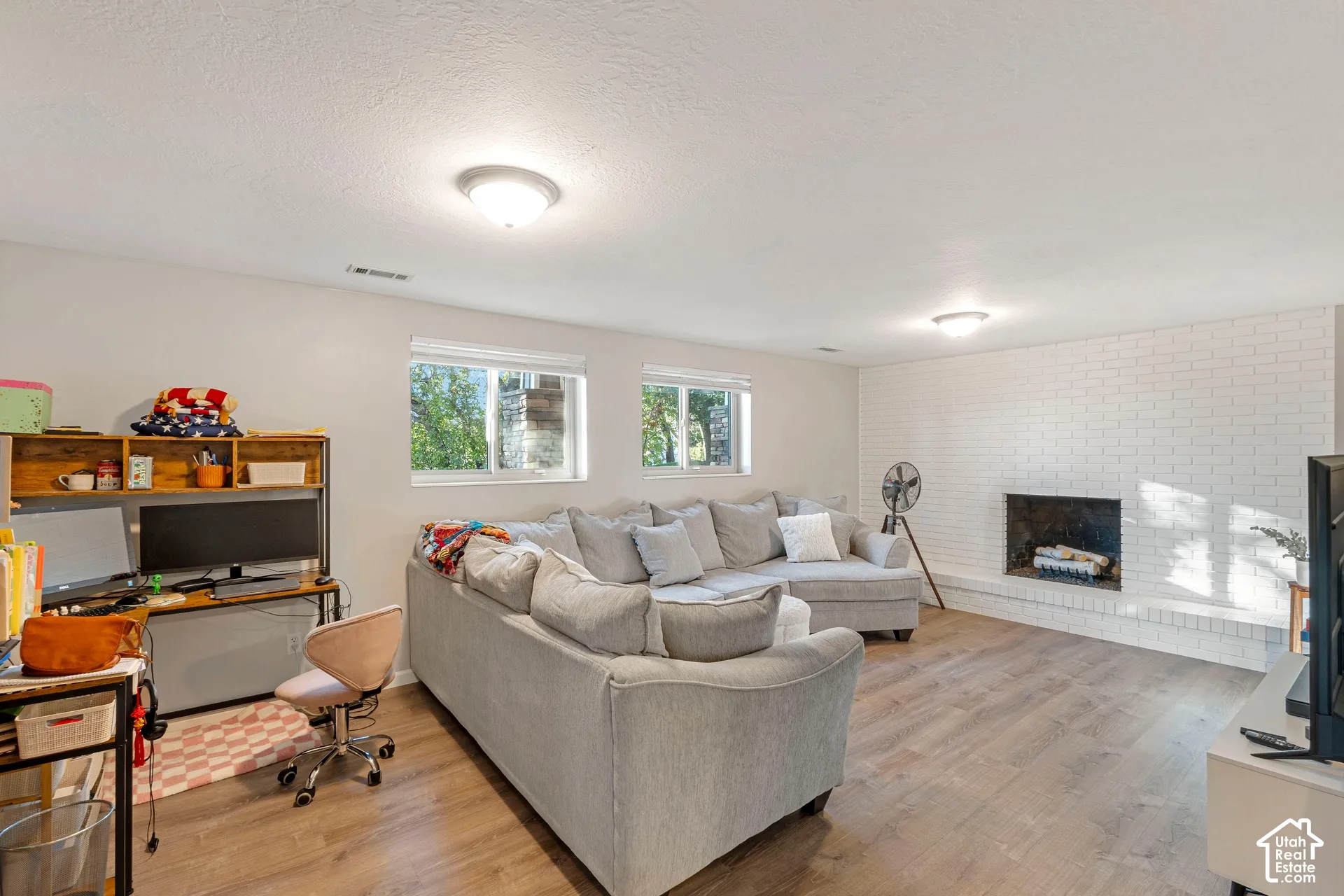 Living area featuring wood-finish flooring, white-painted brick fireplace with an integrated hearth, and dual windows with natural light