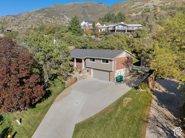 Multi-level residence featuring a brick and vertical siding exterior, an attached two-car garage, stone hardscaping, mature trees, and a mountain backdrop