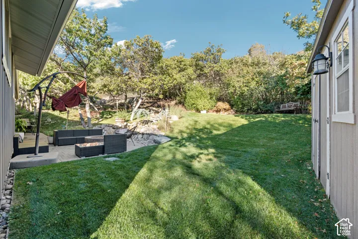 Backyard featuring a paved stone patio, built-in rock landscape elements, mature trees, and a detached storage structure with a wall-mounted light fixture