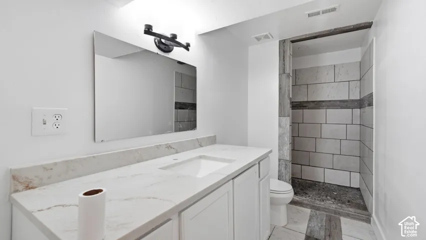 Bathroom featuring a vanity with a white countertop and integrated sink, a rectangular wall mirror, matte black vanity lighting, and a walk-in shower with textured tile surround and stone-style flooring