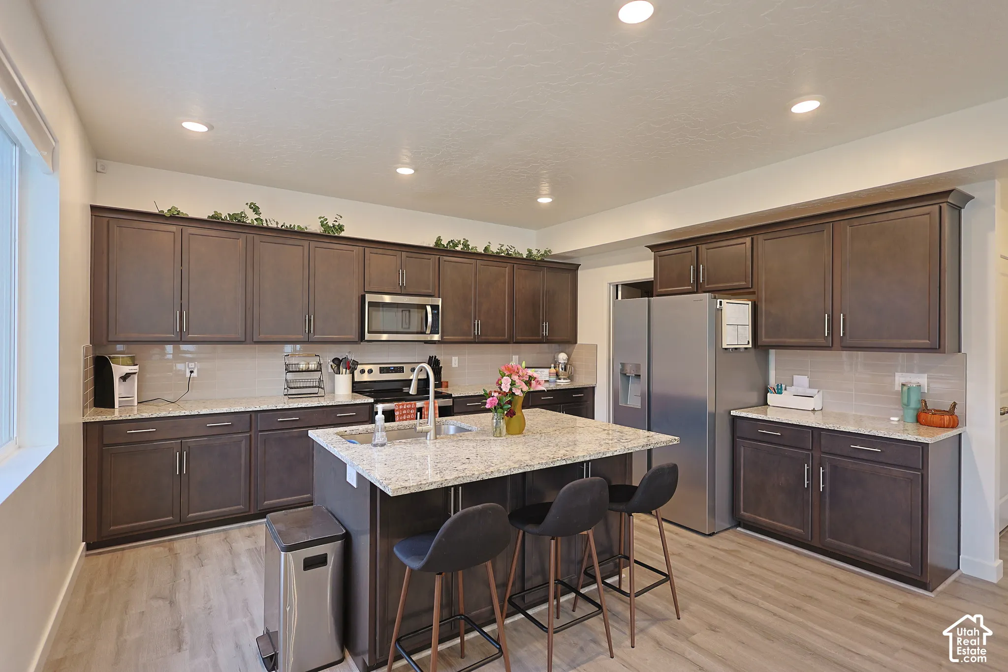 Kitchen featuring dark-finish cabinetry, stone-surface countertops, recessed ceiling lighting, a central island with an integrated sink, and wood-finish flooring